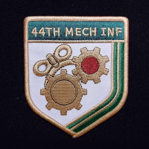 May include: A white patch with a green border and gold stitching. The patch features a gold gear design with a red centre and the text "44TH MECH INF" in black.