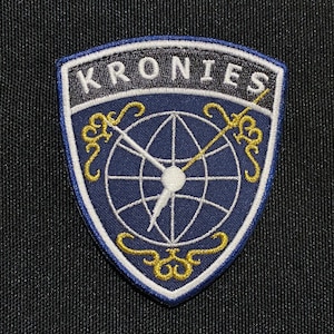 May include: A blue and white embroidered patch with the text "KRONIES" in white and a stylised globe design with a white compass needle. The globe is surrounded by a gold-coloured design.