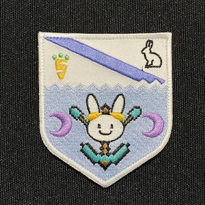 Op de afbeelding: Een witte, blauwe en paarse geborduurde patch met een cartoonkonijn dat een geel hoofdband draagt en een wortel vasthoudt. Het konijn wordt omringd door twee halve manen. De patch heeft een blauwe diagonale streep en een witte achtergrond.