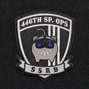 Puede incluir: Un parche bordado en blanco y negro con el texto "446TH SP. OPS" y "SSRB" en una pancarta debajo. Un animal de dibujos animados gris con ojos azules y una antena amarilla está en el centro del parche.