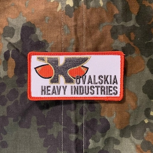 May include: A rectangular patch with a white background and an orange border. The patch features a stylized black and orange logo with the text "OVALSKIA HEAVY INDUSTRIES" in black.