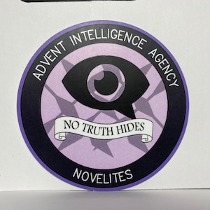 Op de afbeelding: Een ronde sticker in paars en zwart met de tekst "Advent Intelligence Agency" en "No Truth Hides" in een banner. De sticker heeft ook een gestileerde oogafbeelding en de tekst "Novelites" onderaan.