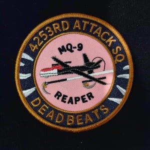 Op de afbeelding: Een geborduurde patch in roze en blauw met de tekst "4253rd Attack Sq Deadbeats" en "MQ-9 Reaper" met een zwart-wit silhouet van een drone.