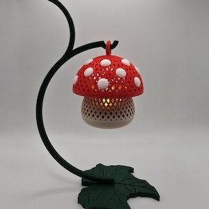 May include: A whimsical mushroom-shaped lamp with a red cap and white polka dots. The lamp has a beige base and sits on a dark green, curved stand with a leaf-shaped base. The lamp is designed to hold a small light.