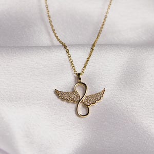 May include: A gold necklace with a pendant shaped like an infinity symbol with wings. The wings are covered in small, clear stones.