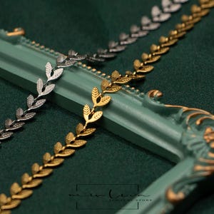 May include: Close-up of three leaf-shaped metal chains in silver, gold, and gunmetal colors. The chains are arranged diagonally across a teal-colored decorative frame with gold accents, set against a dark green fabric background. The chains are likely for jewelry making.
