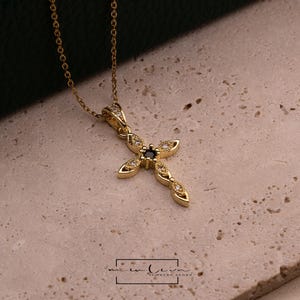 May include: A gold-colored cross pendant necklace with a small, dark blue center stone and clear accent stones. The cross is on a delicate gold chain. The necklace is displayed on a textured, light-colored surface.
