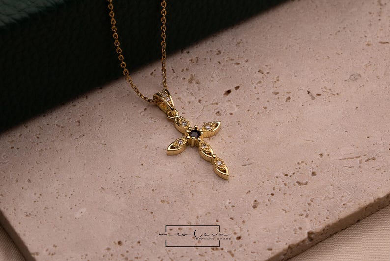 May include: A gold-colored cross pendant necklace with a small, dark blue center stone and clear accent stones. The cross is on a delicate gold chain. The necklace is displayed on a textured, light-colored surface.