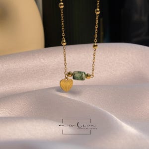 May include: A gold chain necklace with a green stone bead and a heart pendant. The necklace is on a white fabric background.