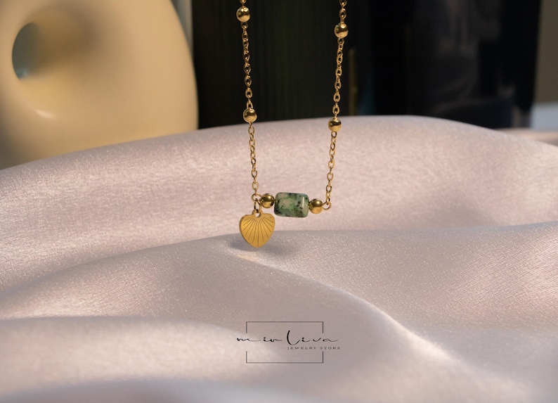 May include: A gold chain necklace with a green stone bead and a heart pendant. The necklace is on a white fabric background.