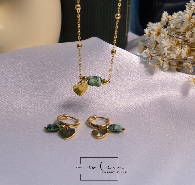 May include: A gold chain necklace with a green stone pendant and a heart charm. Matching earrings with green stones and heart charms are also included. The jewellery is displayed on a white surface.