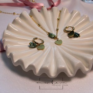 May include: A gold necklace and earrings set with green stone and leaf charms. The jewellery is displayed on a white ceramic dish with a scalloped edge.