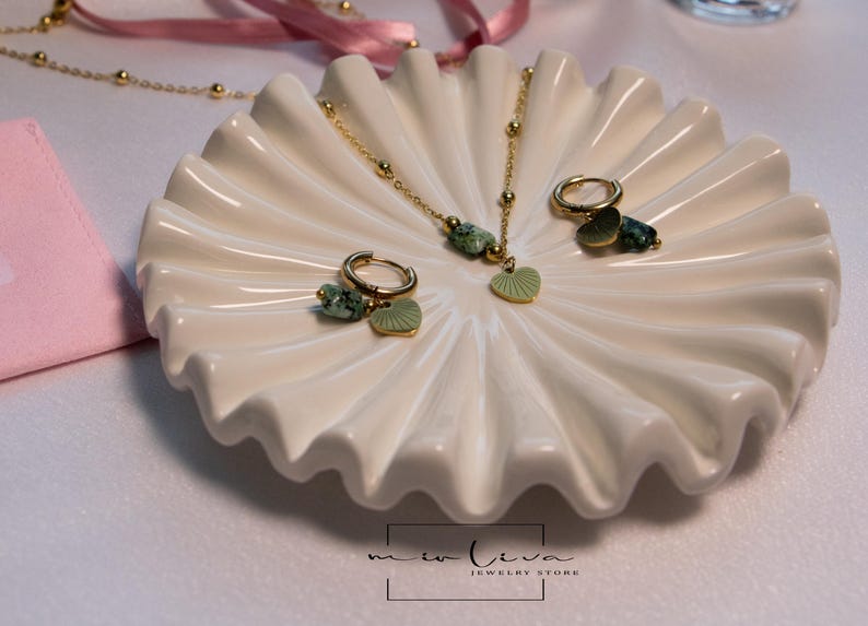 May include: A gold necklace and earrings set with green stone and leaf charms. The jewellery is displayed on a white ceramic dish with a scalloped edge.