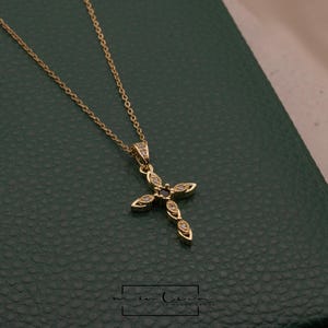 May include: A gold-colored necklace with a cross pendant. The cross is adorned with small, clear gemstones and a dark center stone. The necklace is displayed on a dark green surface.