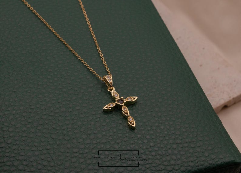 May include: A gold-colored necklace with a cross pendant. The cross is adorned with small, clear gemstones and a dark center stone. The necklace is displayed on a dark green surface.