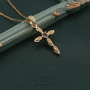 May include: A gold-colored cross pendant necklace with a delicate chain. The cross is adorned with small, sparkling gemstones and a central dark stone. The necklace rests on a green surface.