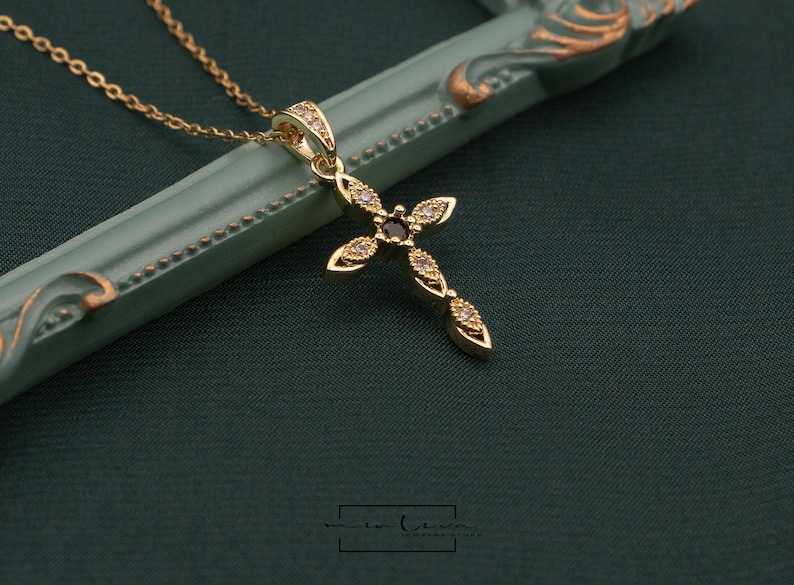 May include: A gold-colored cross pendant necklace with a delicate chain. The cross is adorned with small, sparkling gemstones and a central dark stone. The necklace rests on a green surface.