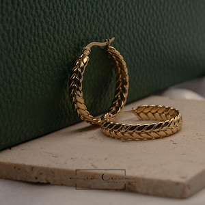 Peut inclure: Une paire de créoles dorées avec un motif tressé. Les boucles d'oreilles sont présentées sur une surface claire avec un fond vert foncé. Les boucles d'oreilles sont de taille moyenne et ont un fermoir sûr.