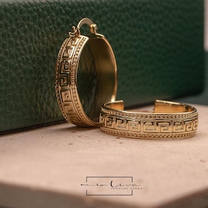May include: A pair of gold hoop earrings featuring a raised Greek key pattern bordered by beaded edges. One earring stands upright while the other lies flat. The text "me liva JEWELRY STORE" is printed in a black rectangular frame at the bottom.