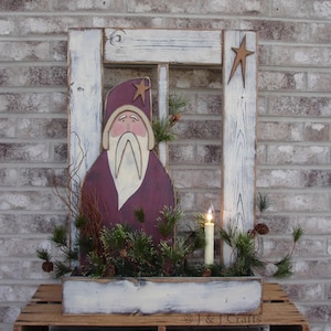 May include: A wooden window frame decoration featuring a painted figure of a bearded man in a purple robe and hat. The frame is distressed white with two wooden stars, green pine branches, small pinecones, and a single electric candle light.