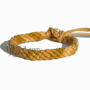 May include: A braided hemp bracelet with a brown and yellow color gradient. The bracelet has a small white bead in the center.