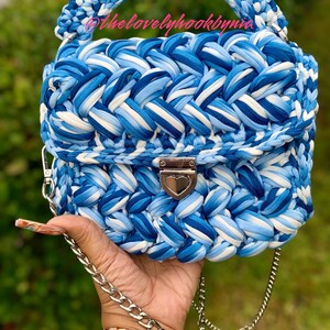 May include: A blue and white crocheted handbag with a silver chain strap and a heart-shaped clasp. The bag has a braided design and is made with a thick yarn.