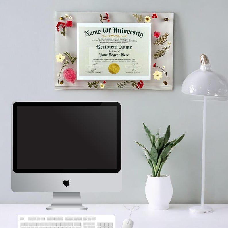 Puede incluir: Un diploma en un marco acr&iacute;lico transparente, adornado con flores secas de colores. El diploma tiene el texto "Name of University", "Recipient Name" y "Your Degree Here". El marco est&aacute; sobre un escritorio blanco con una computadora, planta y l&aacute;mpara.