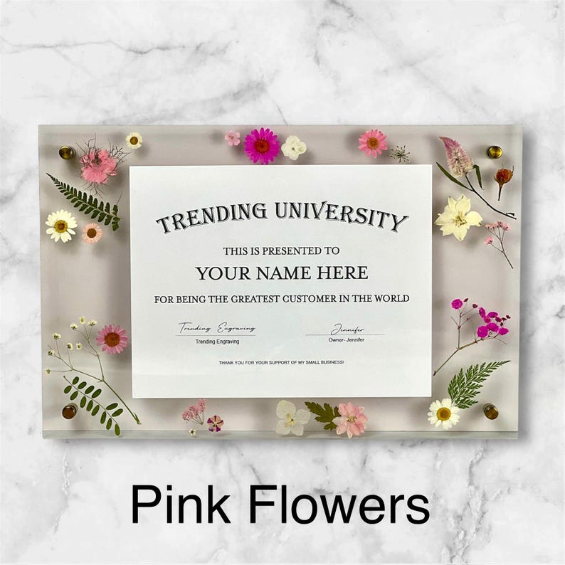 Puede incluir: Un marco de acr&iacute;lico transparente con un certificado que dice "TRENDING UNIVERSITY" y "YOUR NAME HERE". El marco est&aacute; decorado con flores rosas y blancas y follaje verde. El texto "Pink Flowers" est&aacute; en la parte inferior.