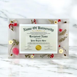 May include: A clear acrylic frame encases a diploma, adorned with dried flowers and foliage. The diploma reads "Name Of University" and "Recipient Name" with the degree information. Gold-colored hardware secures the frame.