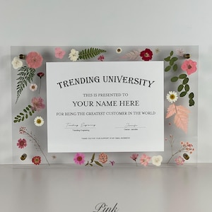May include: A clear acrylic plaque with the text "TRENDING UNIVERSITY" and "YOUR NAME HERE" surrounded by pink and white dried flowers and greenery. The plaque includes the text "FOR BEING THE GREATEST CUSTOMER IN THE WORLD". The word "Pink" is at the bottom.