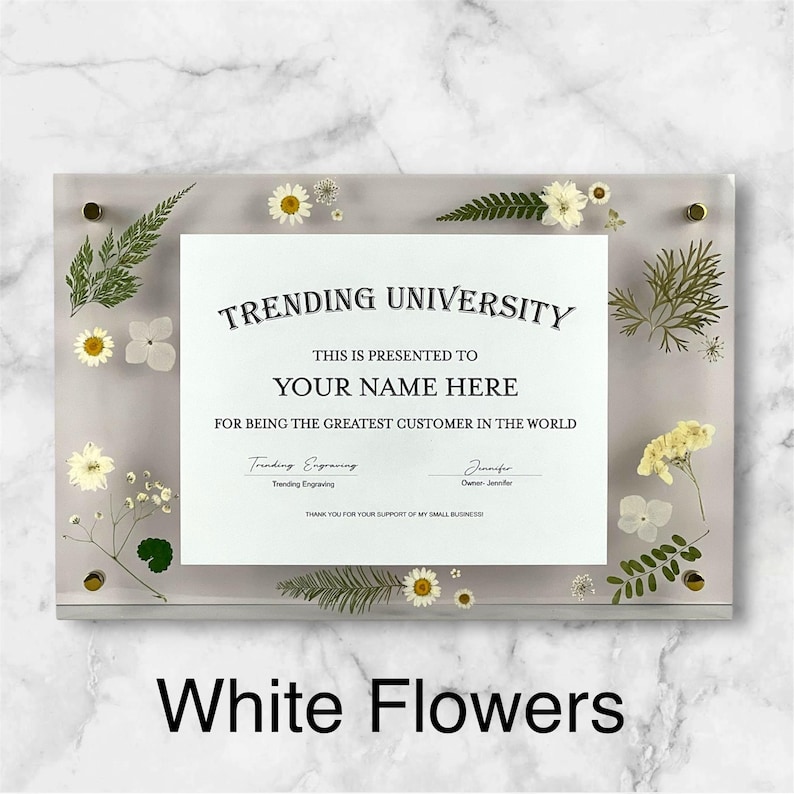 Puede incluir: Un marco rectangular de acr&iacute;lico transparente con un certificado que dice "TRENDING UNIVERSITY" y "YOUR NAME HERE". El marco est&aacute; decorado con flores blancas y vegetaci&oacute;n. El texto "White Flowers" est&aacute; en la parte inferior.