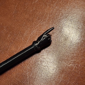 Könnte beinhalten: Ein schwarzer Zauberstab mit einem Zeigefinger. Der Zauberstab zeigt eine Hand mit ausgestrecktem Finger, die an einem langen, zylindrischen Griff befestigt ist. Der Zauberstab besteht aus einem dunklen Material und befindet sich auf einem braunen Lederhintergrund.