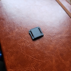 Peut inclure: Un petit clip en plastique noir imprimé en 3D, de forme rectangulaire et à surface texturée. Le clip repose sur un carnet en cuir marron.