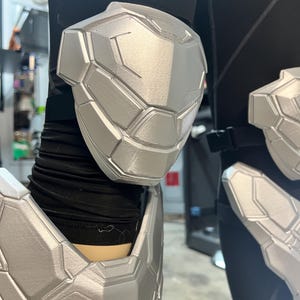 May include: A set of silver-colored, segmented armor pieces worn over black fabric. The armor includes a knee guard with a geometric, multi-paneled design and a matching shin guard, both featuring a smooth, metallic finish and black straps.