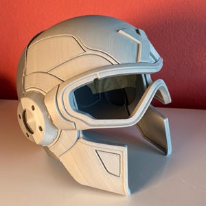 May include: A light gray, textured helmet featuring a prominent brow ridge, a central forehead emblem, and a dark, tinted visor. The helmet includes side circular accents and a structured face guard that extends downward, showcasing a layered design.