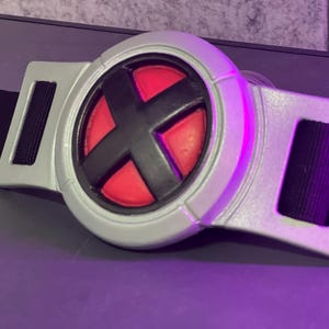 May include: A silver-colored belt buckle with a black and red circular emblem featuring a black "X" design. The buckle is attached to a black fabric belt. The buckle is made of a hard plastic material.
