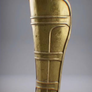 May include: A golden-colored greave, a piece of armor for the lower leg. The metal has a slightly aged appearance with horizontal and vertical decorative lines. The greave is curved to fit the leg.