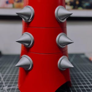 May include: A red cylindrical object with six silver spikes evenly spaced around its exterior. The spikes have a conical shape and are set against a silver circular base. The object has a smooth, glossy finish.