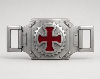 Puede incluir: Una hebilla de cinturón rectangular de metal plateado con un diseño de engranaje central y un emblema de cruz roja oscura. La hebilla tiene un acabado metálico cepillado, remaches visibles y dos bucles rectangulares en los lados para la correa.