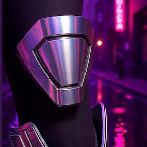 May include: A silver metal arm guard with a geometric design. The arm guard is worn on a black sleeve. The background is a blurred street scene with neon lights.