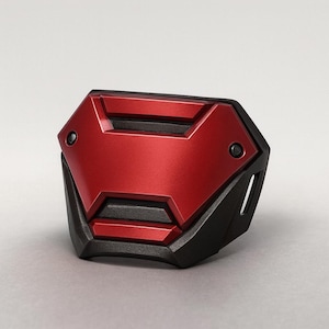 May include: A red and black geometric object with a metallic finish. The object has a central red panel with a textured surface, framed by black angular edges. Two small black circular details are visible.