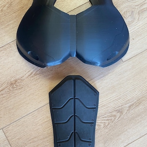 May include: Black 3D-printed armor pieces. The set includes a chest plate and a back plate. The chest plate has a curved design with two rounded sections. The back plate is rectangular with curved indentations and small circular details.