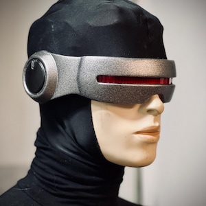 May include: A futuristic silver and red visor with a black head covering. The visor has a sleek, metallic design with a red horizontal light. The head covering is made of a black, form-fitting material. The item is displayed on a mannequin.