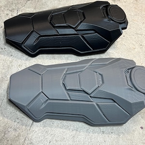 May include: Two forearm guards featuring a segmented, geometric armor design. One guard is matte black, and the other is matte gray. Each piece has a raised circular detail at the top and multiple angular panels across the surface, creating a structured, protective appearance for the arm.