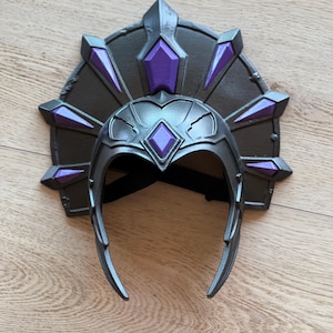 May include: A dark gray headpiece featuring a fan-shaped back crest and a front frame, both adorned with multiple pointed purple diamond-shaped accents. The structure includes a central diamond detail on the forehead and a black elastic strap.