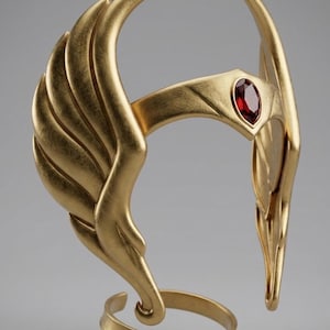 May include: A gold-colored headpiece featuring large, stylized wings on each side that curve upward. The front of the band displays a central, marquise-cut deep red gemstone set within a pointed gold frame. The base includes a circular gold band.
