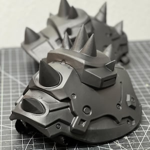 May include: A pair of matte black shoulder pauldrons featuring multiple sharp, conical spikes and molded geometric paneling. The armor pieces are constructed with rigid, angular sections and include integrated black straps for secure attachment.