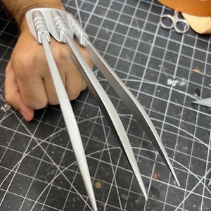 Può includere: Un accessorio da mano color argento caratterizzato da tre lunghe estensioni appuntite a forma di lama fissate su una piastra di base. L'oggetto con finitura metallica è progettato per essere indossato sul dorso della mano, con ogni lama che si estende verso l'esterno.