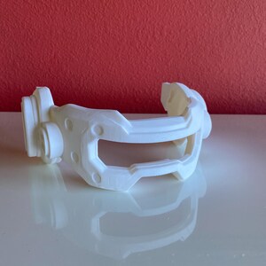 Puede incluir: Un componente de auriculares blanco impreso en 3D con un diseño futurista. El componente presenta una diadema curva, una placa frontal con una abertura y piezas laterales circulares. El objeto está sobre un fondo rojo.