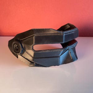 May include: A black, 3D-printed mask with a geometric, angular design. The mask features a horizontal eye opening, a prominent circular detail on the side, and a layered, rigid structure that covers the upper face area with sharp, defined edges.